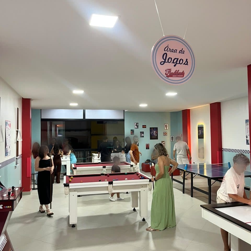 O salão cheio à noite, com a plaquinha "Área de Jogos" no teto, mesas de sinuca, ping-pong, hockey de mesa e fliperama, e gente jogando